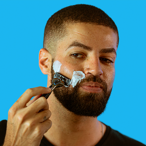 Um homem de cabelo raspado curto segura o aparelho de barbear contra a bochecha, que está coberta de espuma.