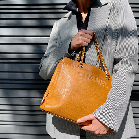 Mulher vestindo um blazer cinza segura uma bolsa de couro da marca Chanel na cor caramelo. A foto mostra apenas o torso da mulher, que está diante de um painel cinza.