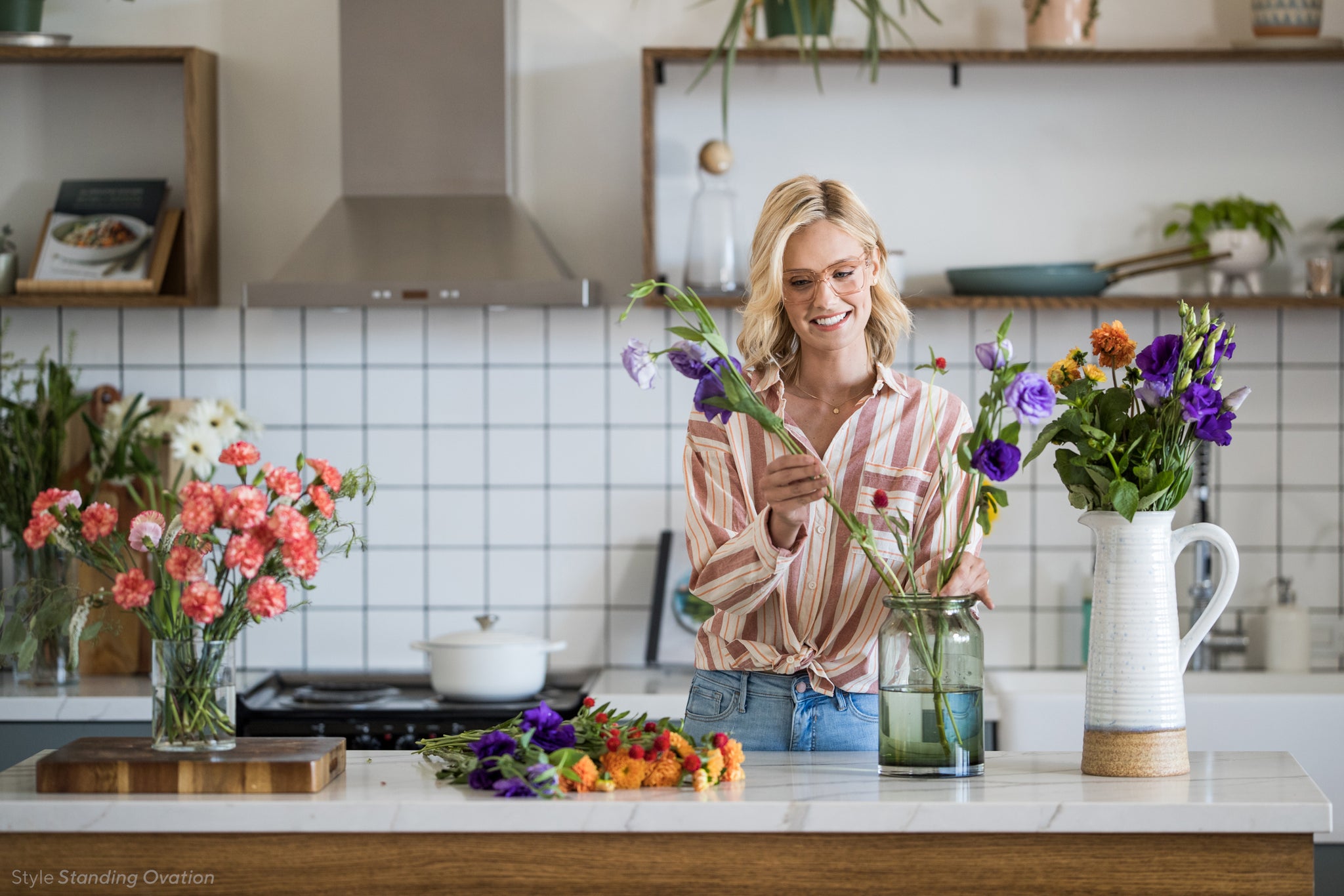 Blonde woman arranging flowers in her kitchen and wearing Peepers Standing Ovation blue light reading glasses