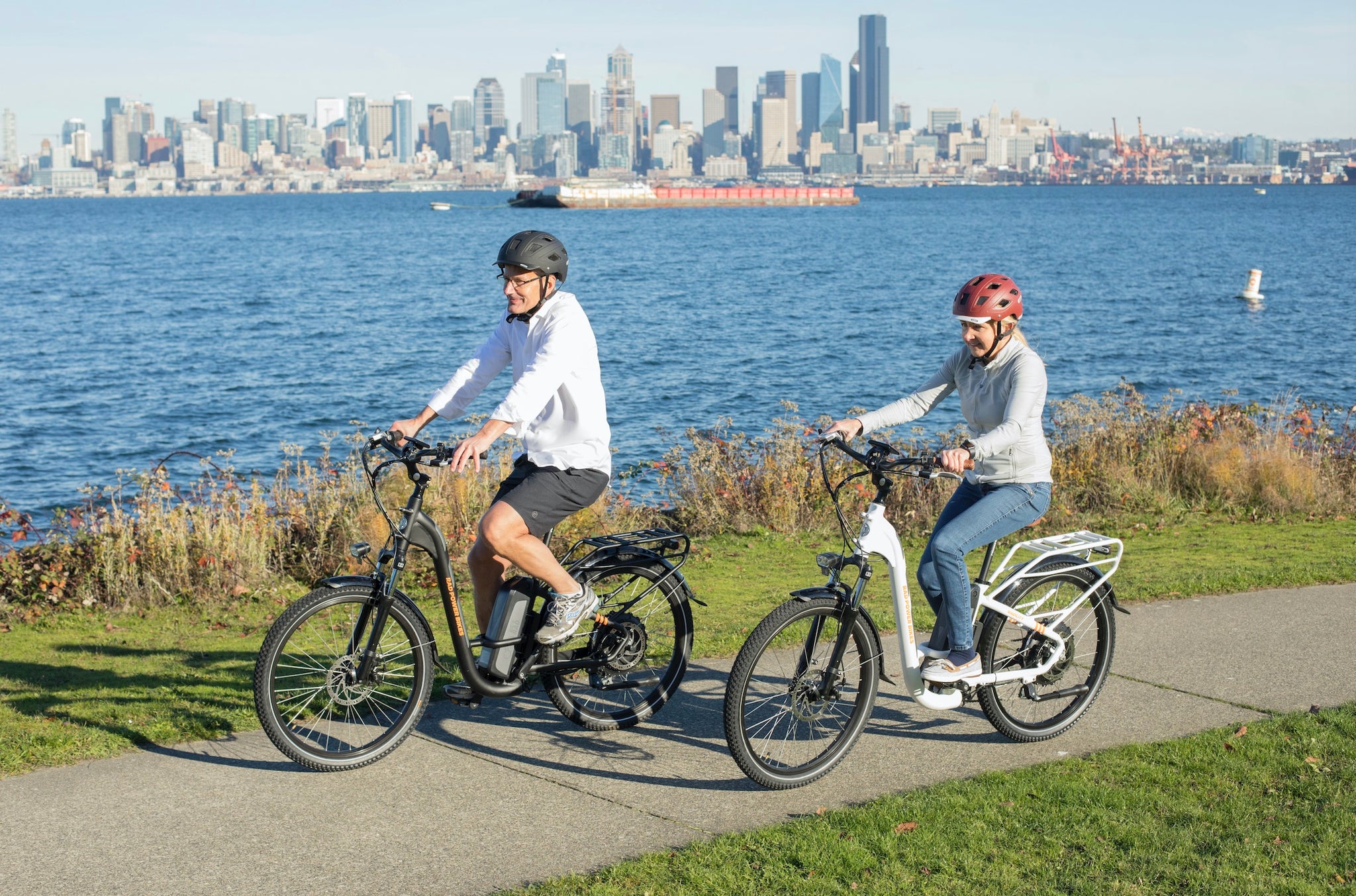 A couple rides a pair of Rad City electric bikes along the waterway. A city skyline is in the background.