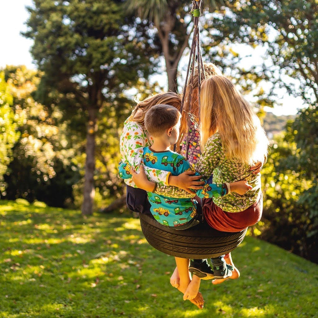 Four kids swing on a tire over a grassy backyard, dressed in colourful clothes from manitoulinsturtlecreek, and in bare feet. The late afternoon sunlight is dappled by trees..