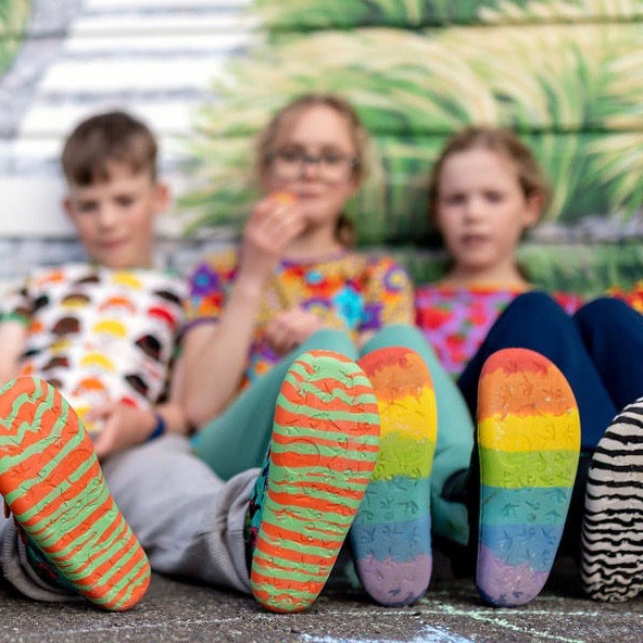 Young person sits on a skatepark rail, the camera focuses on her white PaperKrane rainbow sneakers