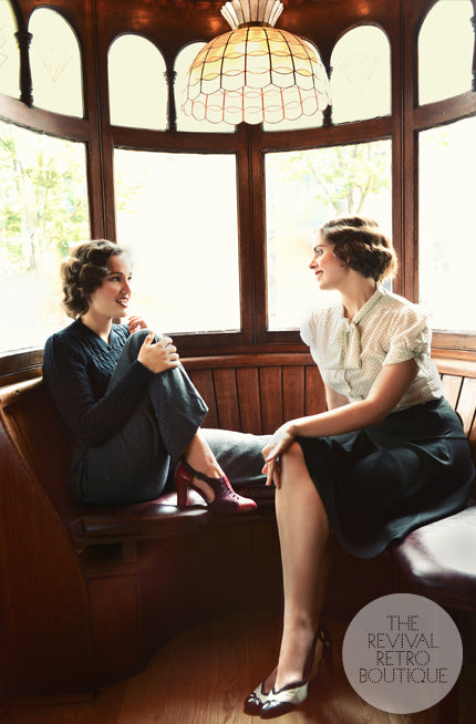 Two women sit in an old fashioned window seat, their clothing is 1940s inspired and both wear statement vintage shoes.