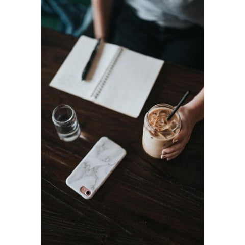 Person sitting at a wooden desk with their notepad, pen and iced coffee in a glass with a paper straw