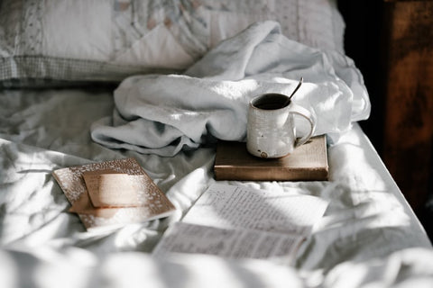 A cosy set up in a bed with a mug of coffee, a book and white cotton bed sheets 