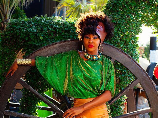 model showcasing flowing african headpiece, tribal green shirt and orange pants against wooden wagon wheel