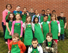 A group of students and their teacher in front of a brick wall. They are all wearing green aprons.