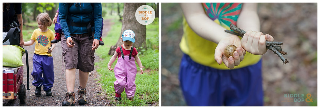 two kids in rainpants hiking next to mother's legs, mom wearing baby in carrier on her back. | child's hands with seed pods.