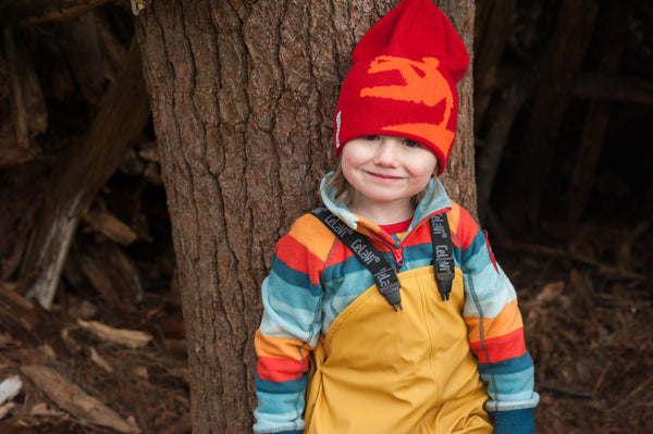 child wearing red hat, multi colored fleece, and golden yellow bibbed rainpants, standing in front of a tree.