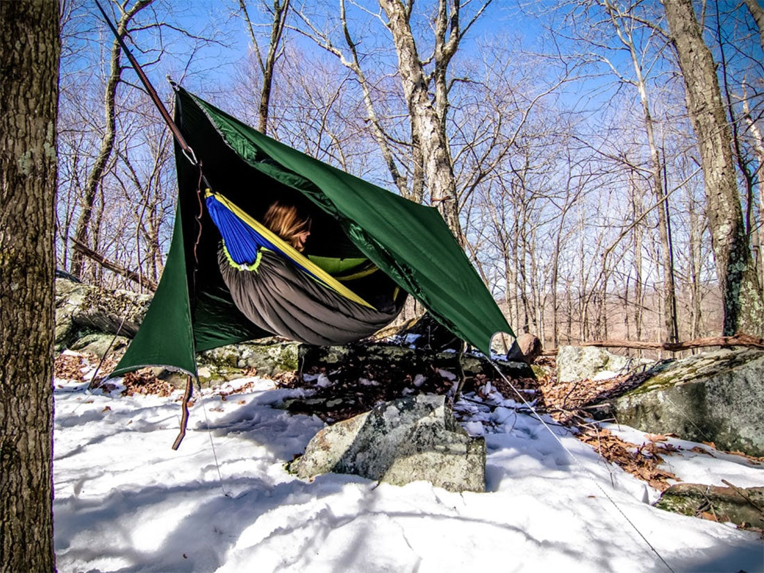 Camper laying in their hammock with a rain cover in the snowy outdoors.