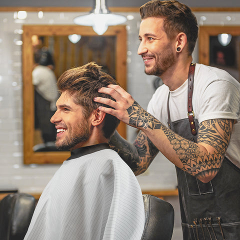tattooed hairdresser cutting mans hair while wearing a neocape
