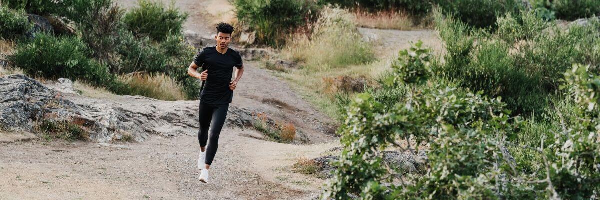 A runner facing the camera at a slight angle to the right, running on sand with green grass behind him. IT Band Syndrome, ITBS Running, Runners Knee, knee braces running, running with ITBS