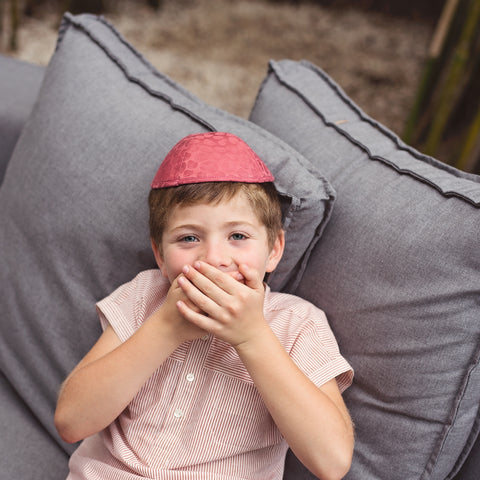 A boy laughing while sitting on a grey couch wearing a red iKIPPAH.