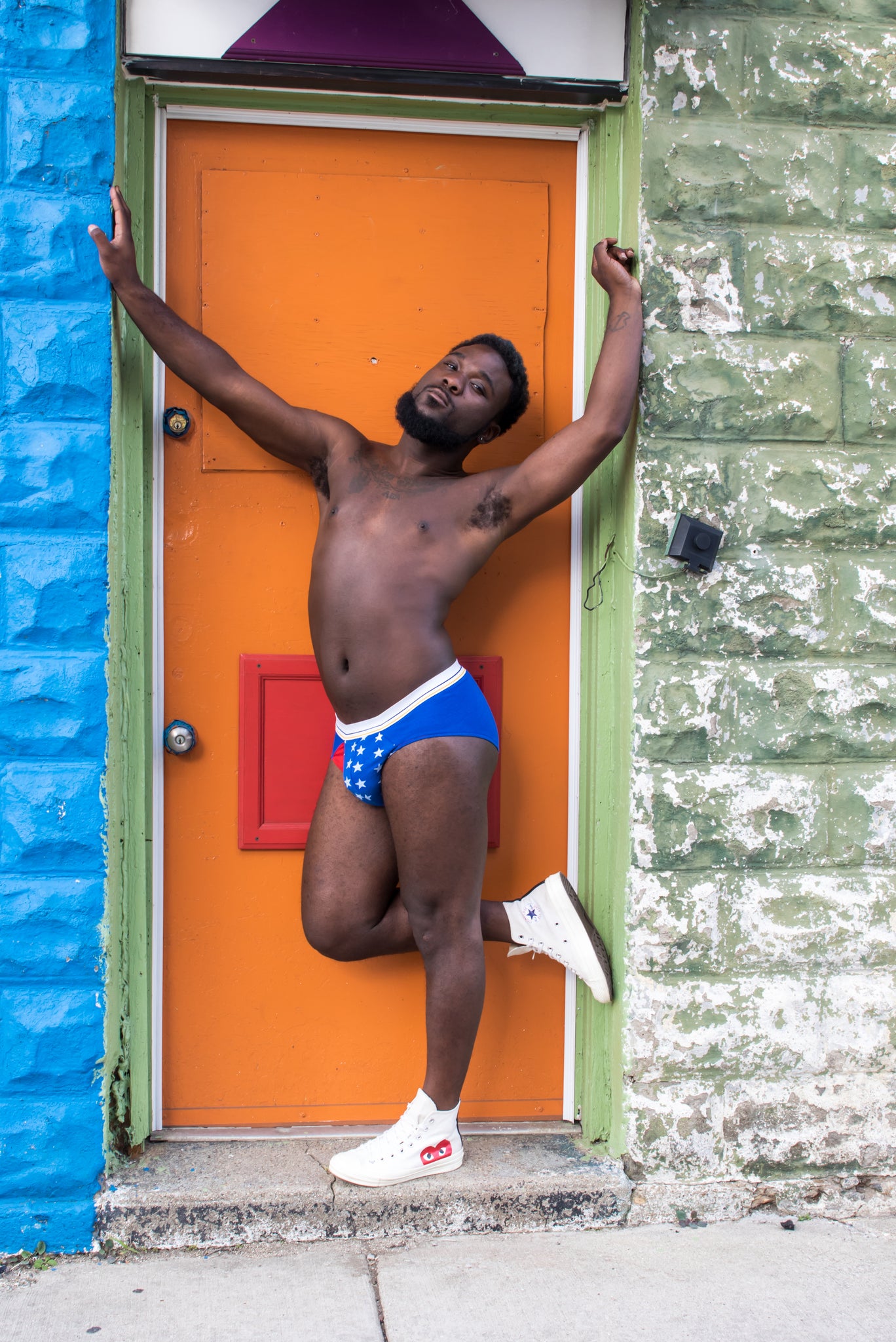 An underwear clad man standing in an orange doorway. His undies are blue with white stars, and a red pop of color at the fly.