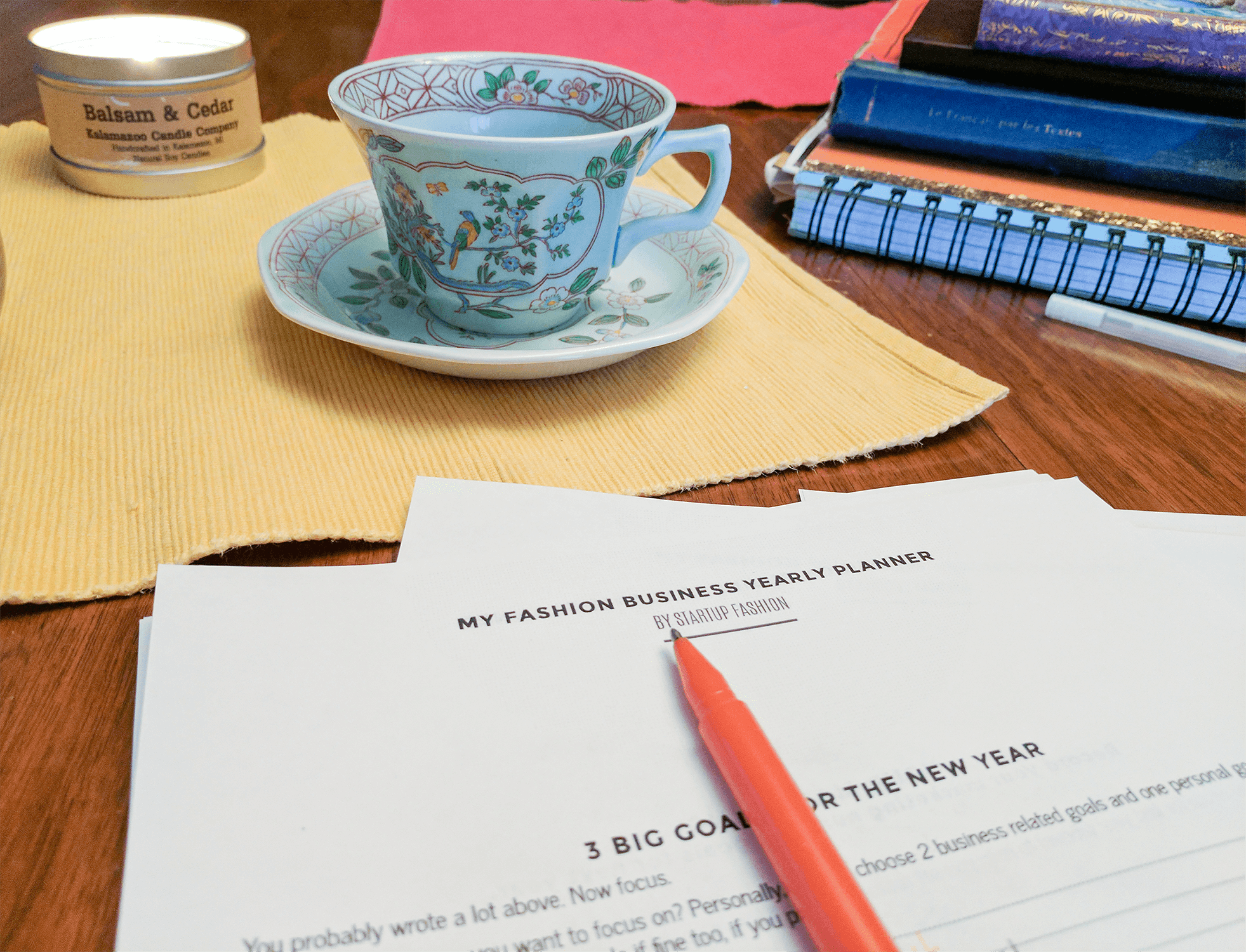 A table spread with notebooks, a beautiful old tea cup, and a burning candle. Startup Fashion's yearly planner is in the foreground with an orange pen.