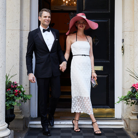 Bride wearing Jane Summers strapless white lace tea length going away dress with a pink hat with black feathers and groom wearing a black tuxedo