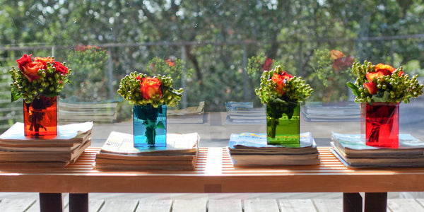 Flowers arranged in four 105C boxes of various colors, sitting on a bench in front of a window