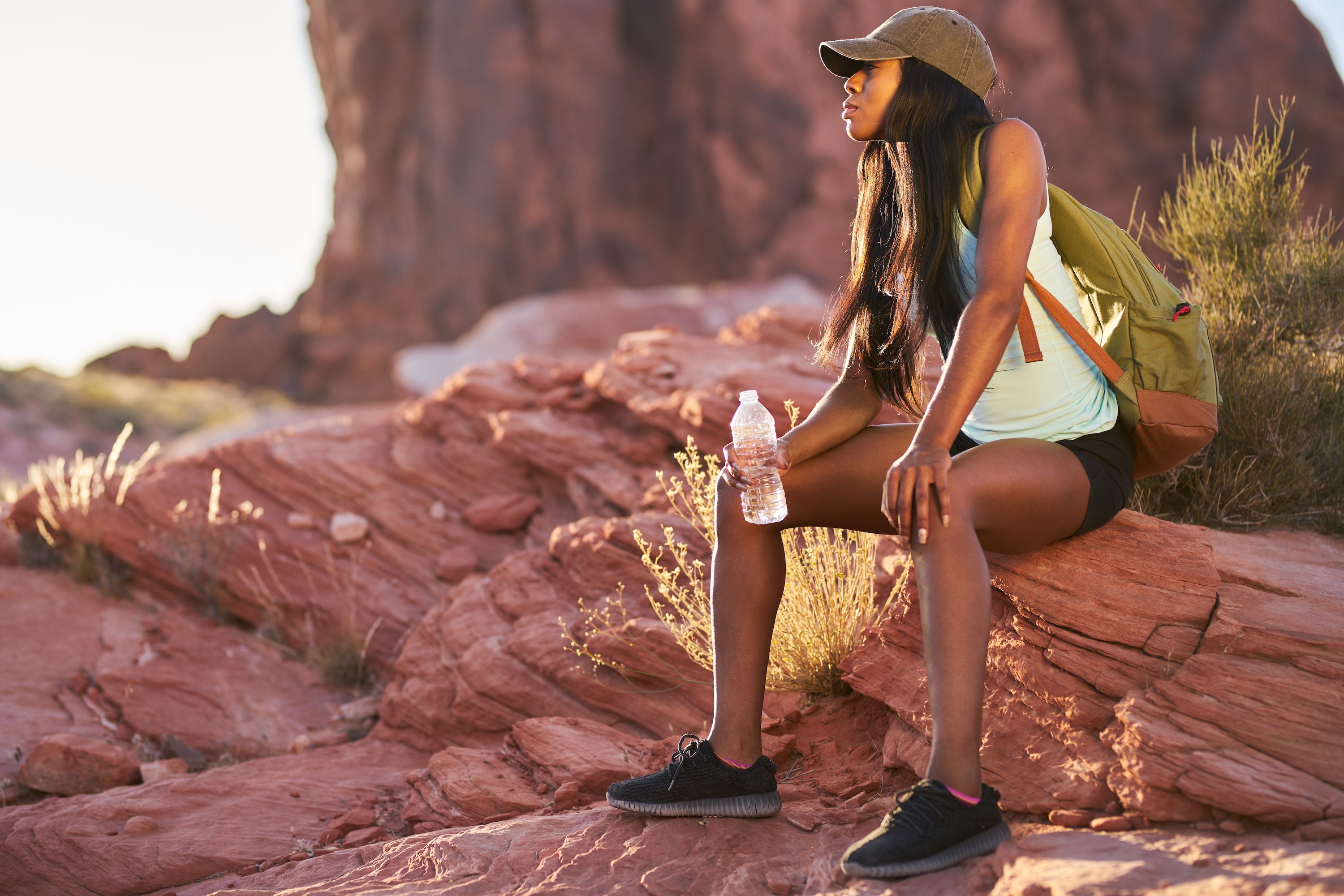 african american girl with hat and backpack sitting on red rock drinking water