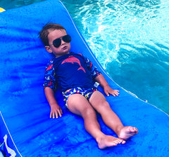 little boy sunbathing on a raft in a pool wearing sunglasses and BBLittles Shark Rashguard and swim diaper