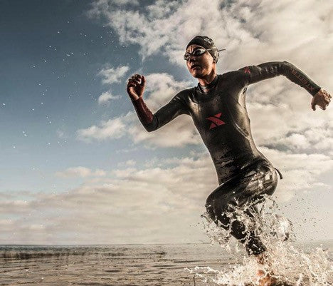 A female triathlete wearing a full-length wetsuit made by Xterra exits the ocean. Xterra wetsuits are featured in the Mother's Day gift guide by H2O Audio. 