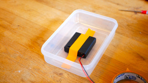 Image of a black battery box taped down inside a Tupperware container with orange tape