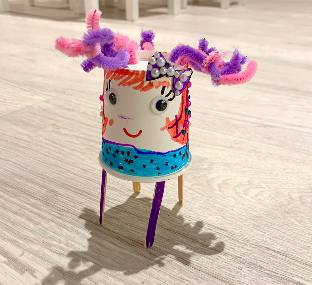 Paper cup based robot with face with two goggly eyes, drawn on eyelashes and pink hair, a light blue body with black spots, plus pink purple pipe cleaner hair and four purple coffee stirrer legs  