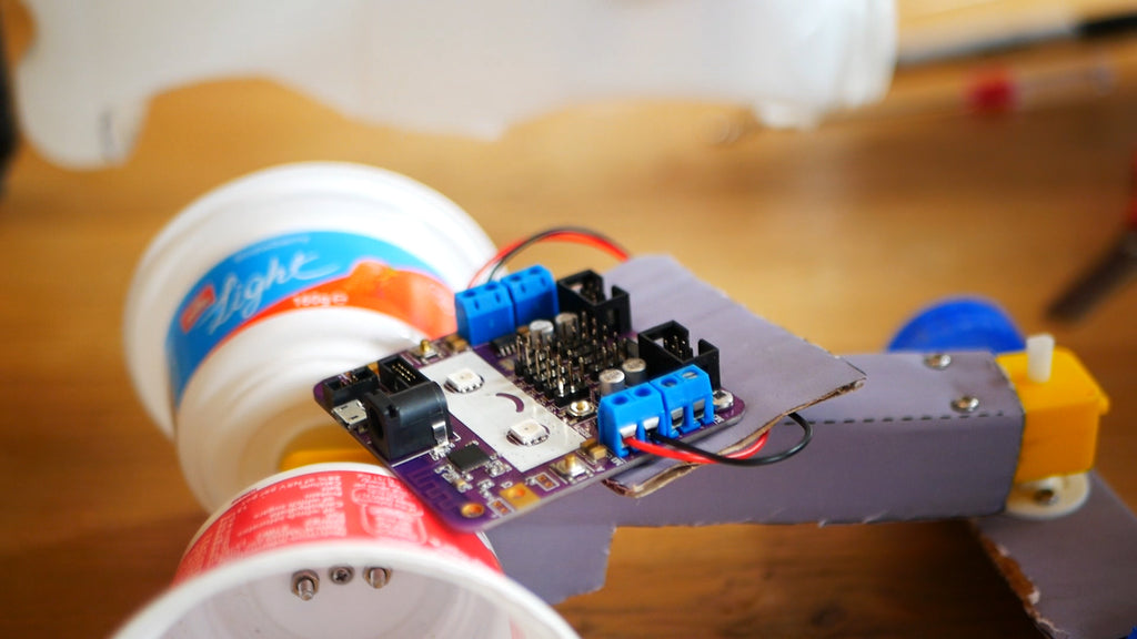 Image of a purple circuit board with a smiley face connected to black and red wires, laying on a cardboard structure.
