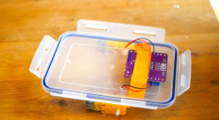 Image of a purple circuit board taped on a Tupperware container lid with orange tape