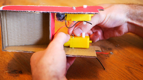 Image of a yellow motor being screwed on to a red cardboard structure.