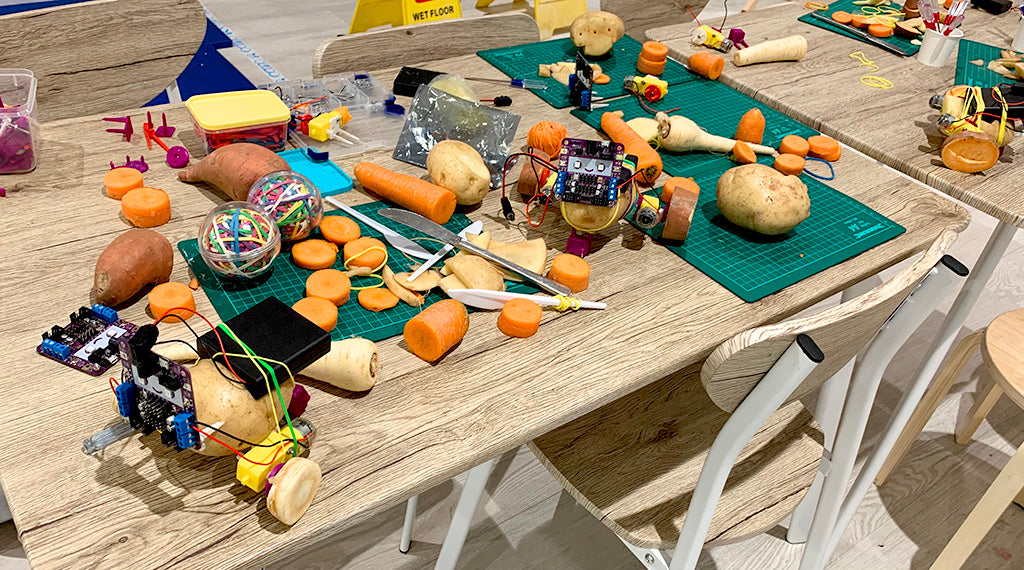 Photo of a messy table covered with potato, carrots and parsnips, electronics and tools, plus three potato robots.
