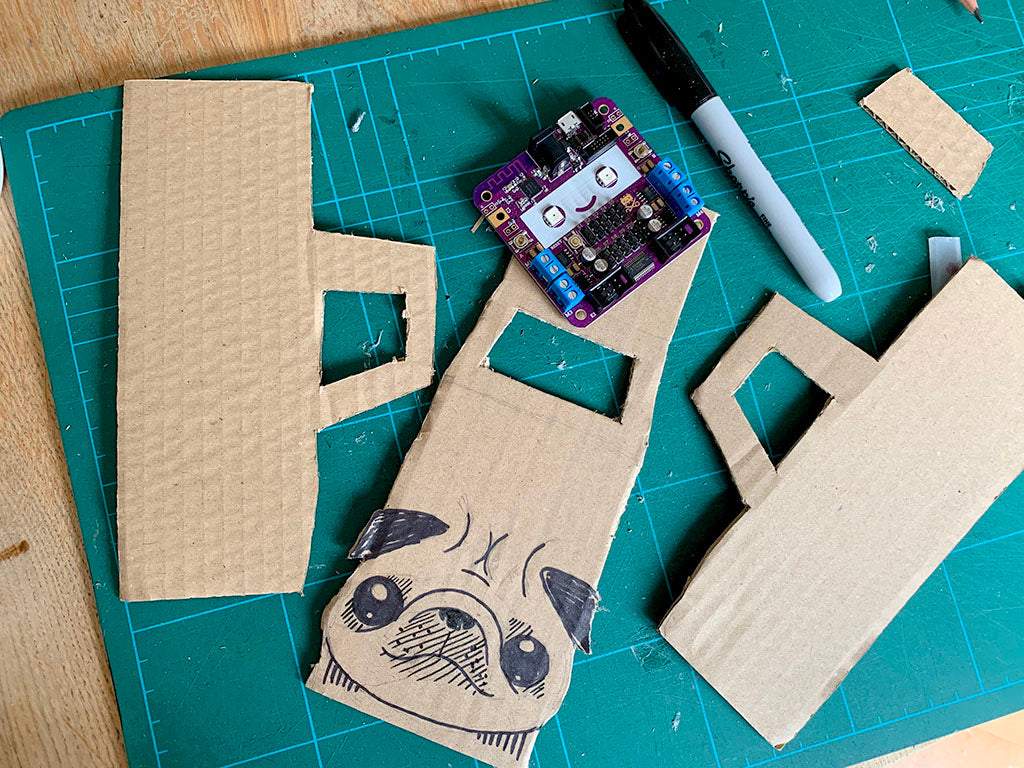 Photo of some cardboard monster truck parts, a purple smiling circuit bard and a black marker pen on a cutting mat