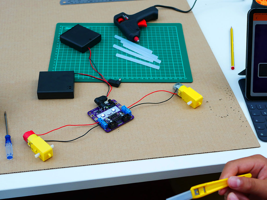 Photo of a Smartibot circuit board connected to two motors and a battery box, lying on a large sheet of cardboard. In the background a cutting mat, glue gun and hot melt glue sticks