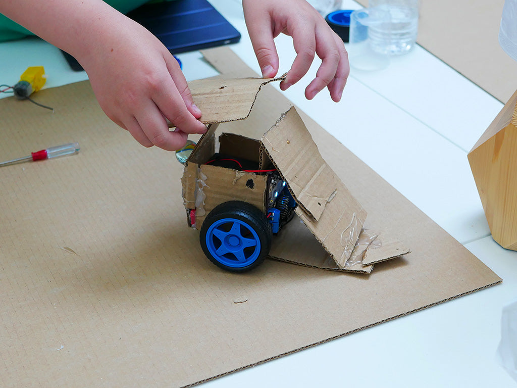 Photo of a cardboard robot chassis like in the example to which a young person is adding extra cardboard parts to form a narrow wedge.