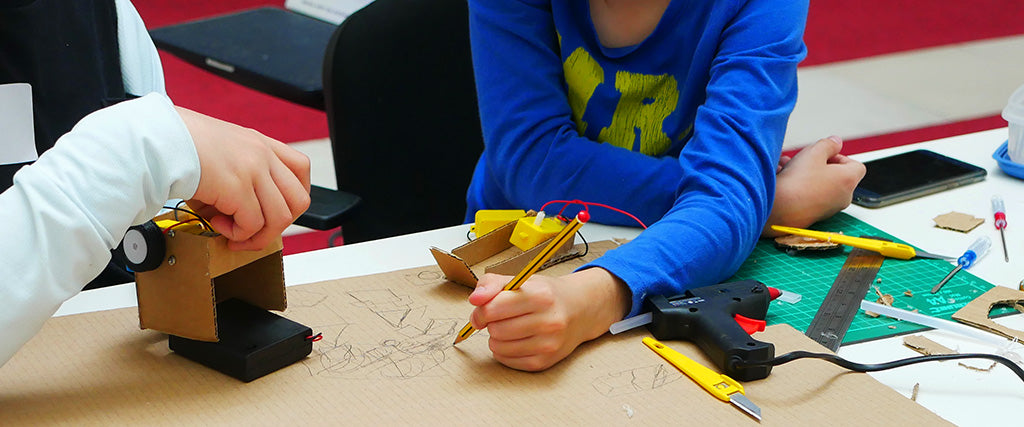 Photo of two young people collaborating to build a cardboard robot. One sketches in pencil on a piece of cardboard whilst another holds a partially completed robot. 