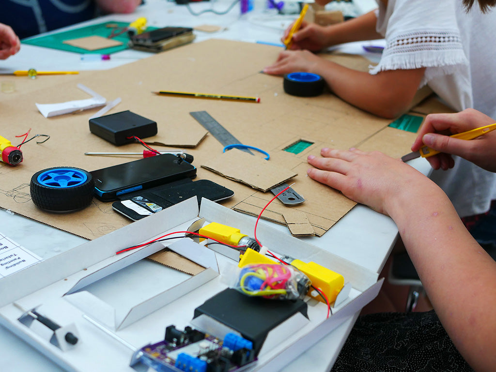 Photo of two young people cutting large sheets of cardboard with craft knives. Parts from a Smartibot kit lie in the foreground.