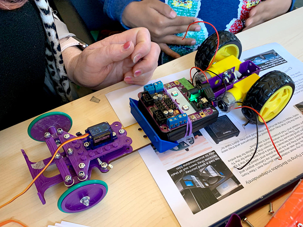 Photo of a small car chassis with a purple plastic steering mechanism and wheels at the front, two yellow plastic motors and wheels at the back and a smiling purple circuit board in the centre
