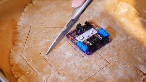 Image of a piece of gingerbread dough, with a purple circuit board next to it, and a silver knife cutting the dough.