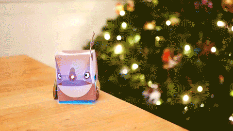 A GIF of a reindeer robot with a light-up nose, driving around with a christmas tree in the background.