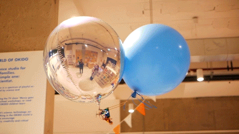 A GIF of a blimp, made out of two helium-filled balloons and powered by a purple circuit board, hovering around an office. 