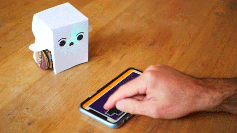 A looping video of a white paper robot being driven using a control pad on a smartphone