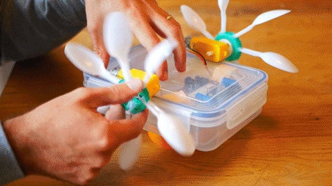 A looping GIF of someone rotating a propeller, which is made from a green milk bottle lid and plastic spoons, attached to a yellow motor