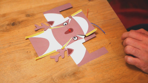 Image of a paper reindeer laying flat on a wooden table, with pieces of double sided tape stuck to it.