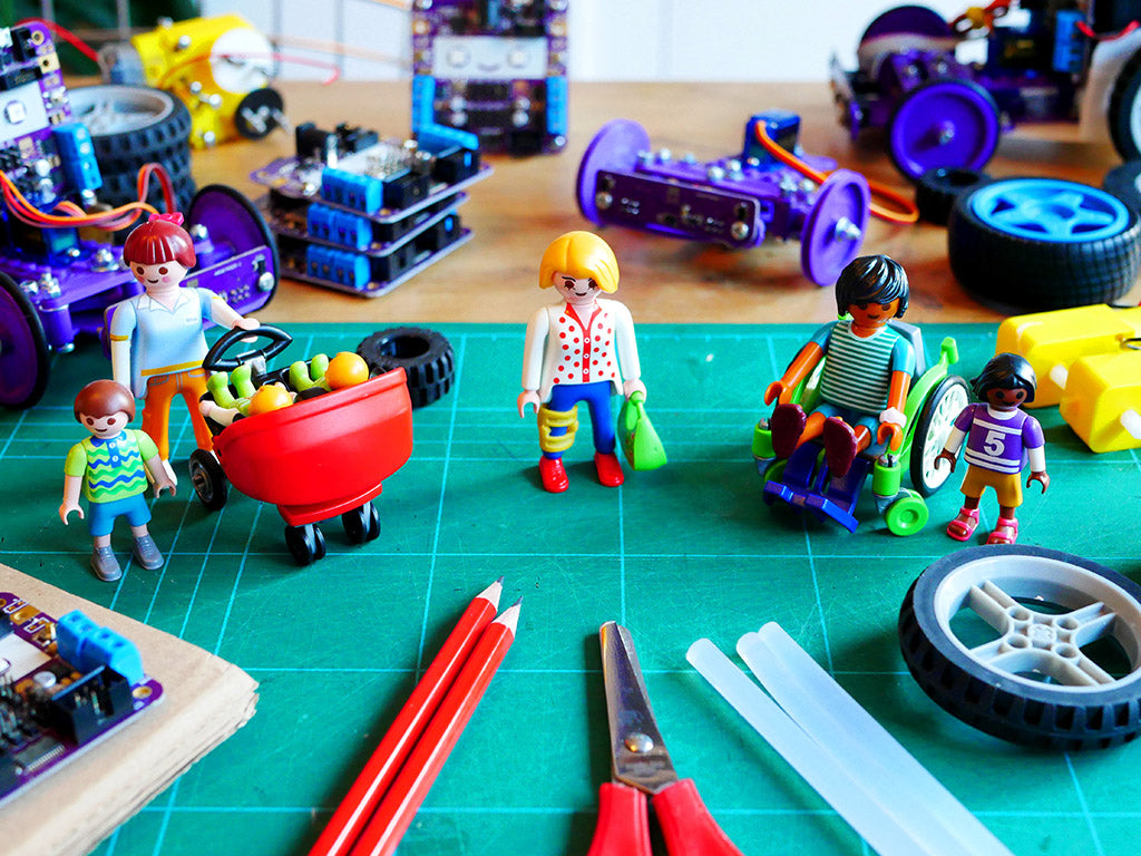 Photo of some Playmobil plastic figures, one of a woman with a buggy with two babies in and and a small child, one of a woman wearing a leg brace and one of a man using a wheelchair with a small child. They are standing on a cutting matt and surrounded by purple circuit boards, craft materials and tools and small wheels and motors.  