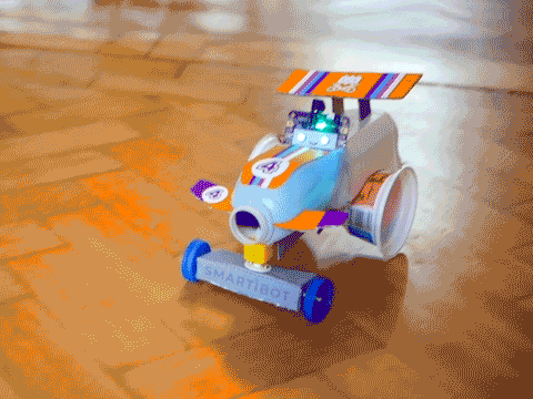 Looping video of a milk-bottle bodies, yoghurt pot wheeled car, with a smiling circuit board in the drivers seat, driving back and forward on a wooden floor
