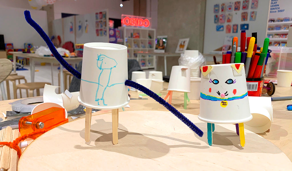 Two paper-cup based robots made by children, one with long blue pipe cleaner arms and a blue figure drawn on the front, the other decorated to look like a cat 
