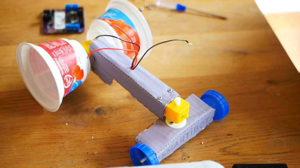 Image of a race car made from a cardboard axle and chassis, two yoghurt pots as rear wheels and two blue milk bottle lids as front wheels.