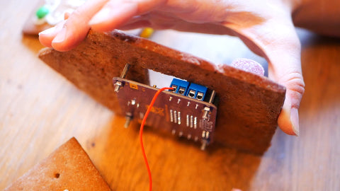 Image of a circuit board attached to a piece of gingerbread house, and a red wire attached to the circuit board.