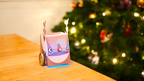 Image of a paper reindeer and a red light-up nose, with a Christmas tree in the background.