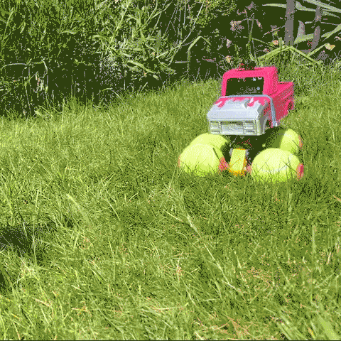 Looping video of a pink toy monster truck with yellow tennis ball wheels driving over grass