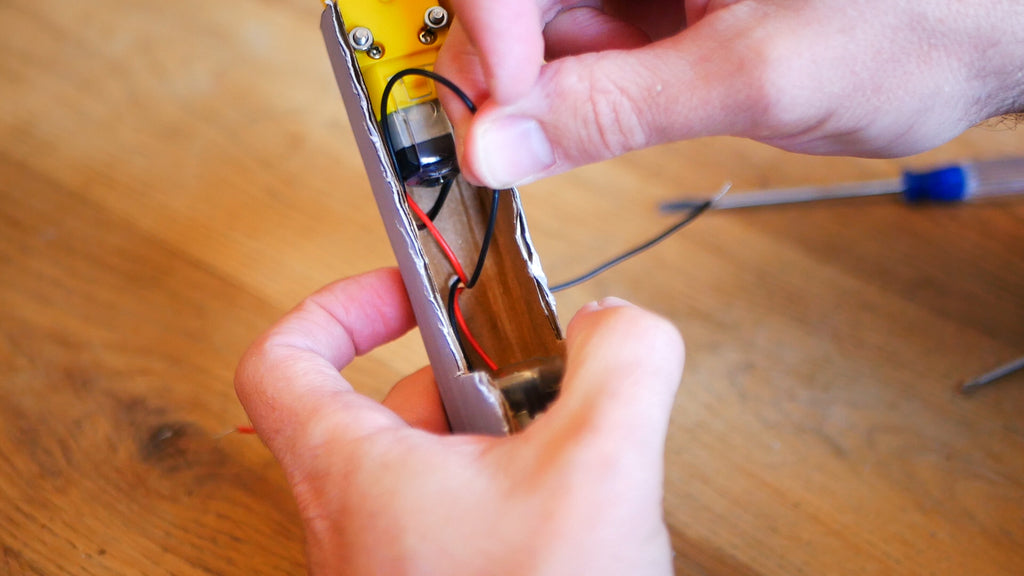 Image of a cardboard front axle and two yellow motors, where the red and black wire of the upper motor are being inserted into a small hole in the cardboard.